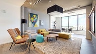 This contemporary lounge space makes an immediate impression with its bold, eclectic design and thoughtfully curated interiors. The single-floor layout opens with a welcoming entry featuring large glass doors, porcelain tile flooring, and a wall-mounted flat-screen TV set into a warm walnut wood surround — a striking focal point that sets the tone for the rest of the space.

The main lounge area is defined by a dramatic black-and-white houndstooth accent wall, against which a large, vibrant portrait painting adds a pop of color and artistic energy. Seating is arranged in a relaxed, social configuration, combining modular sectional sofas in muted gray and olive tones, a wood-veneer lounge chair, and a teal accent ottoman, all anchored by a textured area rug and an oval walnut coffee table.

Floor-to-ceiling windows along one wall flood the space with abundant natural light and offer expansive views of the surrounding cityscape and skyline. A built-in bistro-style seating nook with a small round table and modern armchairs sits directly beside the windows, ideal for casual meetings or quiet moments.

Overhead, the ceiling features track lighting, recessed fixtures, and a bold black pendant element that adds architectural dimension. The overall ambiance is modern, vibrant, and inviting — a space that balances comfort with contemporary design flair.