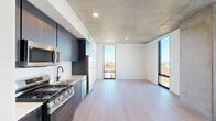Located at 625 North 2nd Avenue in downtown Phoenix, AZ, this modern single-floor unit offers a thoughtfully designed open layout that maximizes every square foot. The combined living, dining, and kitchen area serves as the heart of the space, characterized by striking exposed concrete ceilings and light wood-tone flooring that runs throughout. Floor-to-ceiling windows on multiple walls flood the interior with abundant natural light and frame sweeping views of the surrounding Phoenix cityscape and beyond, creating an airy, elevated feel. Exposed concrete structural columns add an industrial-chic aesthetic that complements the clean, contemporary design. The kitchen is fully equipped with stainless steel appliances — including a gas range, built-in microwave, dishwasher, and refrigerator — dark cabinetry extending to the ceiling, light quartz countertops, and a white tile backsplash. The entrance hall leads to a dedicated laundry area, a closet for storage, and a full bathroom. The bathroom features large-format gray tile flooring, a sleek floating vanity with an integrated sink, a backlit mirror, matte black hardware accents, and a glass-enclosed tub and shower combination. The overall ambiance is clean, modern, and urban, making this a well-appointed space in the heart of Phoenix.