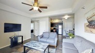 This single-floor residence offers a thoughtfully designed open-concept layout that seamlessly connects the main living, dining, and kitchen areas. The living room is centered around a comfortable seating arrangement and features dark hardwood-style flooring that runs throughout the space, adding warmth and visual continuity. A ceiling fan with integrated lighting fixture provides both comfort and illumination, while a tray ceiling detail adds an architectural touch to the room.

The kitchen is equipped with stainless steel appliances, including a refrigerator and microwave, and is complemented by dark cabinetry and track lighting overhead, creating a sleek, modern aesthetic. The open sightlines between the kitchen and living area enhance the sense of spaciousness and make the layout ideal for everyday living and entertaining.

Adjacent to the living room, a dedicated dining area offers space for a full dining set, maintaining the open flow of the floor plan. Multiple doorways lead to additional rooms within the unit, suggesting private spaces such as bedrooms and bathrooms tucked away from the main living areas. The overall design palette — featuring neutral gray tones, dark flooring, and white trim — creates a cohesive, contemporary ambiance throughout the space.
