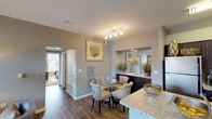 This single-floor residence offers a thoughtfully designed layout that balances comfort and style throughout. The open-concept kitchen is equipped with dark espresso cabinetry, stainless steel appliances, and a granite-topped peninsula with bar seating — ideal for casual dining or entertaining. Adjacent to the kitchen, the dining area features hardwood-style flooring and track lighting overhead, creating a warm and inviting atmosphere. A built-in sideboard with a granite surface and mirrored backdrop adds a refined touch to the dining space. The living room is generously sized, bathed in natural light from a large sliding glass door that opens onto a private balcony. Rich hardwood-style floors, a cozy fireplace, and bold wall art contribute to the room's welcoming character. Two bedrooms provide comfortable retreats; the primary bedroom includes plush carpeting, a ceiling fan, large windows, and direct access to an en-suite bathroom. Both bathrooms are finished with dark wood vanities, granite countertops, and hardwood-style flooring. The second bathroom features a glass-enclosed shower, while the primary bath offers a spacious vanity setup. Neutral gray tones and warm gold accents are carried consistently throughout, lending the entire space a cohesive and polished aesthetic.