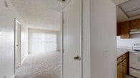 This single-floor, one-bedroom, one-bathroom unit delivers a practical and efficient layout across a comfortable footprint. The combined kitchen and living room serves as the heart of the home, with the living area featuring plush wall-to-wall carpeting, a ceiling fan with light fixture, and a sliding glass door flanked by vertical blinds that allow generous natural light to fill the space. The kitchen is equipped with warm oak-toned cabinetry, light marble-patterned countertops, a coil-burner electric range with overhead ventilation, and a sink, providing ample workspace and storage for everyday cooking needs. The bedroom is a well-proportioned retreat with soft carpeting underfoot and two large windows dressed with vertical blinds, creating a bright and airy atmosphere. The bathroom features a vanity with an integrated sink, light countertop surfaces, and a wall-mounted mirror. A hallway connects the main living areas to the bedroom and bathroom, adding a sense of separation and privacy. Throughout the unit, textured ceilings, neutral wall tones, and clean white trim contribute to a cohesive, understated aesthetic that is easy to personalize.