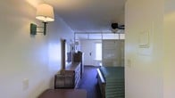 Located at 1865 Lakeland Dr in Jackson, Mississippi, this single-floor room offers a straightforward and functional layout designed for comfortable short-term stays. The main sleeping area is anchored by a king-sized bed dressed in a green coverlet, positioned beneath a ceiling fan with an integrated light fixture that helps maintain a comfortable atmosphere throughout the room. A full-length dresser with a large mirror sits along one wall, providing ample storage and a practical vanity area. Wall-mounted sconce lighting adds supplemental illumination near the entry side of the room. The space features dark carpeting throughout, lending a cozy, grounded feel to the interior. A partially open partition wall separates the sleeping area from a small foyer near the entrance, where natural light filters in through a window with horizontal blinds adjacent to the front door. The bathroom is accessible from this transitional area, rounding out the practical amenities of the room. The overall design is clean and utilitarian, with a neutral color palette on the walls that keeps the space feeling open and uncluttered. This room is well-suited for travelers seeking essential comforts in a convenient Jackson, MS location.