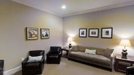 This single-floor space showcases a thoughtfully designed living area that combines comfort with understated sophistication. The room is finished with soft, neutral gray walls that create a calm and inviting atmosphere, complemented by crisp white crown molding that adds a refined architectural detail along the ceiling perimeter. Recessed lighting overhead provides even illumination throughout the space, while a classic table lamp with a warm-toned shade introduces a cozy, ambient glow in the corner.

The seating arrangement is both functional and stylish, featuring a tufted taupe sofa dressed with decorative pillows alongside two dark leather armchairs, each accented with light-colored cushions. A small side table nestled between the seating pieces provides a practical surface for the lamp and additional items. The flooring consists of neutral-toned carpeting that adds warmth underfoot and ties the room's color palette together cohesively.

A gallery-style display of framed black-and-white and sepia-toned photographs lines the walls, lending a curated, artistic character to the space. Overall, the room presents a balanced and polished environment suitable for relaxed lounging or casual conversation.