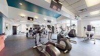 Located at 101 Frankfort Square in Columbus, Ohio, this fitness center offers a well-equipped, single-floor facility designed to support a comprehensive workout experience. The space is defined by its open layout, durable rubber flooring, and a striking teal accent ceiling that adds a bold visual element to the otherwise clean, white interior.

The cardio zone is generously stocked with multiple treadmills, elliptical trainers, and stair climbers, many positioned near large windows that provide ample natural light and views of the surrounding neighborhood. Ceiling-mounted flat-screen televisions are positioned throughout the cardio area, allowing users to stay entertained during their workouts.

The strength training section is equally well-appointed, featuring a full dumbbell rack with a wide weight range, adjustable benches, cable machines, a Smith machine, and additional weight training apparatus. A dedicated wall-mounted information board keeps members informed of relevant facility details.

Near the entrance, a warm orange built-in shelving unit adds a charming architectural touch and provides a resource station for members. The facility is accessed through double glass doors, contributing to an open and welcoming atmosphere. Overall, this fitness center combines functional design with a vibrant aesthetic, creating an inviting environment well-suited for a wide range of fitness activities.