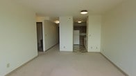 This single-floor unit offers a straightforward, functional layout with clearly defined living and kitchen areas. The main living space features neutral carpeting and cream-colored walls, creating a clean and understated backdrop suitable for a variety of furnishing styles. Flush-mount ceiling light fixtures provide consistent overhead illumination throughout. A structural column near the center of the space subtly divides the living area while maintaining an open feel.

The kitchen, accessible from the main living area, is equipped with dark wood cabinetry, a white refrigerator, and a range. The flooring transitions to a checkered tile pattern, distinguishing the kitchen zone from the carpeted living space. Counter space runs along the wall, providing a practical work area. A hallway extends from the living space, leading toward additional rooms, including what appears to be a bathroom visible in the background. The corridor is lit by a wall-mounted fixture, ensuring adequate visibility. Overall, the unit presents a practical and modest living environment with a neutral, move-in-ready aesthetic.