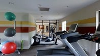 This fitness center is designed to provide a functional and energizing workout environment. The room is outfitted with modern cardio equipment, including Freemotion treadmills and elliptical machines, arranged to make efficient use of the available floor space. The walls feature bold horizontal stripes in shades of yellow, red, and sage green, adding a dynamic visual element that contributes to an active atmosphere. Recessed lighting in the white ceiling ensures the space is well-lit, while a window along one wall introduces natural light. A standout architectural feature is the sleek black-framed glass partition wall, which separates the fitness area from an adjacent space while maintaining a sense of openness and visual connectivity. The flooring combines carpet and hard surface sections, delineating workout zones within the room. The overall design balances practicality with an intentional aesthetic, making the fitness center both a visually appealing and purpose-driven space suited for daily exercise.