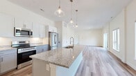 Located at 1471 US-9 in Halfmoon, NY, this thoughtfully designed one-bedroom, two-bathroom residence offers a bright and contemporary living experience across a single floor. The open-concept kitchen and living area forms the heart of the home, anchored by a generously sized granite-topped island with an undermount sink and pendant lighting overhead. White shaker cabinetry, stainless steel appliances — including a refrigerator, range, and microwave — and matching granite countertops complete the kitchen. Light hardwood-style flooring flows throughout the main living areas, enhancing the airy, modern aesthetic. Large windows flood the living room with natural light, while a sliding glass door opens to a private balcony with outdoor views. The spacious primary bedroom features plush carpeting, a bright window, and access to a walk-in closet equipped with wire shelving. The full bathroom includes a gray vanity with granite countertop, built-in shelving, and a soaking tub. A second bathroom provides a toilet and overhead cabinetry with hardwood-style flooring. Clean white walls and recessed lighting throughout contribute to the home's crisp, move-in-ready ambiance.
