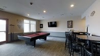 This single-floor game room is designed for leisure and entertainment, offering a well-rounded collection of recreational amenities within a comfortable, open layout. The centerpiece of the space is a full-size pool table with a rich red felt surface, positioned to allow ample movement on all sides. A dartboard mounted on the adjacent wall provides an additional competitive activity, while a wall-mounted flat-screen television serves as a focal point for viewing entertainment. Bar-height seating with dark wood chairs and round tables is arranged along one wall, creating a casual gathering area suited for socializing. Natural light enters through a double-pane window, complementing the recessed ceiling lighting that ensures the room remains well-lit throughout the day and evening. The neutral-toned walls, accented by a lower wainscoting detail, contribute to a clean and polished aesthetic. Carpeted flooring in a textured, low-pile pattern adds comfort underfoot while maintaining a practical surface for the space. Framed wall art adds personality and warmth, completing the inviting, recreational atmosphere of this well-equipped game room.