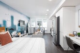 This thoughtfully designed studio unit combines modern aesthetics with practical functionality in a compact, open-plan layout. The space is anchored by a dramatic floor-to-ceiling teal watercolor mural that serves as a bold focal point, adding a vibrant artistic element to the living and sleeping area. Dark hardwood flooring runs throughout, providing a warm contrast to the crisp white walls and ceilings, which are fitted with recessed lighting for a clean, contemporary look. The sleeping area features a wooden-framed bed, while the living space accommodates a sofa, media console with a wall-mounted flat-screen TV, and a dedicated desk nook — ideal for working from home. Natural light filters in through large windows, brightening the space and offering views of the surrounding exterior. The kitchen is well-appointed with stainless steel appliances, including a full-size refrigerator, electric range, over-the-range microwave, and dishwasher, all complemented by light cabinetry, white countertops, and a textured tile backsplash. The bathroom features a sleek vanity with a undermount sink and large mirror, a tiled shower, and the added convenience of an in-unit stacked washer and dryer. Overall, the unit delivers a stylish and highly functional living environment.