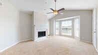 This single-story residence offers a thoughtfully designed layout that balances open living spaces with functional private areas. The living room serves as the centerpiece of the home, showcasing vaulted ceilings that lend a sense of spaciousness and height. A ceiling fan with a light fixture is centrally mounted, and a white-mantled fireplace with a classic tile surround anchors one wall. A projecting bay window arrangement floods the room with natural light and provides views of the surrounding outdoors. An adjacent dining area connects seamlessly to the living room, featuring a pendant light fixture and continuing the neutral carpeted flooring. The kitchen is equipped with light wood cabinetry, dark granite-style countertops, hardwood flooring, and stainless steel appliances including a range, over-the-range microwave, and refrigerator. The hallway leads to the bedrooms, each with soft carpet, a ceiling fan, and a window. The bathrooms feature light wood vanities with granite-style countertops and hardwood flooring. The primary bathroom includes a dual-sink vanity with mirrored closet doors, while a secondary bathroom offers a tub-shower combination. Neutral gray walls and white trim are consistent throughout, creating a cohesive and clean aesthetic.