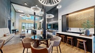 Located at 146 S Wayfinder Ave in Meridian, Idaho, this contemporary two-floor amenity space is defined by its sophisticated architectural design and carefully curated interiors. The ground floor welcomes visitors through wide, wood-floored corridors accented by deep navy and sage green wall treatments, leading into expansive open-concept gathering areas. Soaring double-height ceilings anchor the main lounge, where a bold collection of sculptural, tiered pendant lights serves as a dramatic focal point overhead. Seating areas feature a mix of warm leather sectionals, upholstered accent chairs, and round tables, creating versatile spaces for both casual relaxation and social interaction. A sleek bar-height counter with wood stools runs along one side, complemented by built-in cabinetry and workstation surfaces. Large windows flood the space with natural light and offer views of the surrounding area, while exposed brick columns and textured wall panels add warmth and visual depth. A prominent staircase with metal railings and a wood-capped balustrade connects the two floors, with the upper level featuring an open mezzanine overlooking the main lounge below. Curated abstract artwork adorns the upper walls, enhancing the space's modern aesthetic. Throughout, light hardwood flooring, patterned carpeting, and thoughtful lighting design create a polished, welcoming environment.