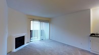 This single-floor residential space presents a straightforward and functional layout designed for comfortable living. The main living area is finished with wall-to-wall carpeting that adds warmth underfoot and contributes to a quiet, subdued atmosphere throughout the room. A classic fireplace is positioned along one wall, serving as a focal point and providing both a practical heating element and an inviting aesthetic anchor to the space. Natural light enters generously through a large sliding glass door fitted with vertical blinds, which offer adjustable privacy and light control. The sliding door opens directly to an exterior area, extending the usable space and providing a seamless connection between indoors and the outdoors. The walls are finished in a neutral, light tone that enhances the brightness of the room and provides a versatile backdrop for a variety of furnishing styles. The overall ambiance is clean, simple, and move-in ready, making it well-suited for those seeking a manageable, low-maintenance living environment. The open feel of the main living area allows for flexible furniture arrangements to suit different lifestyle needs.