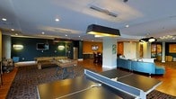 Located at 4451 Telfair Blvd in Camp Springs, Maryland, this dynamic single-floor amenity space is designed to foster entertainment and social interaction. The room is anchored by a variety of recreational offerings, including a ping-pong table, foosball table, and a billiards table, making it a well-equipped destination for leisure activities. Stylish pendant lighting with bold black-and-gold fixtures hangs from the ceiling, complementing the recessed lighting that illuminates the space throughout. The design palette is energetic and modern, with deep teal, rich orange, and dark accent walls that create distinct zones within the open floor plan. Hardwood flooring and patterned carpet help define the different activity areas. A kitchenette or wet bar area is visible in the background, complete with a stainless steel refrigerator and bar-height seating, ideal for casual gatherings. Wall-mounted flat-screen televisions are positioned throughout, adding to the entertainment value of the space. A comfortable lounge area with plush seating provides a relaxed retreat for those looking to unwind. The overall ambiance is lively and inviting, thoughtfully designed to serve as a central hub for recreation and community engagement.