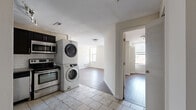 This single-floor, 4-bedroom, 1-bathroom unit offers a practical and well-organized layout across a compact but efficiently utilized footprint. The kitchen is equipped with stainless steel appliances, including a range and over-the-range microwave, dark cabinetry with a tile backsplash, and light tile flooring. A stacked washer and dryer unit is conveniently integrated into the kitchen area, adding in-unit laundry functionality. The full bathroom features tile flooring, a dark-toned vanity with a sink, a standard toilet, and a full soaking tub with a shower combination, finished with clean white surroundings. The four bedrooms vary in size and character — the spacious primary bedroom is carpeted and benefits from multiple windows that bring in generous natural light and outdoor views, creating a bright and airy atmosphere. Additional bedrooms feature either rich hardwood-style flooring or carpet, each with windows providing natural light. The common areas also feature hardwood-style flooring, contributing to a cohesive and clean aesthetic throughout. The neutral color palette of warm whites and beiges across all rooms creates a calm, versatile environment suitable for a variety of furnishing styles.