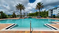 Located at 14050 Integra Dr in Jacksonville, FL, this outdoor amenity space delivers a resort-style experience designed for relaxation and recreation. The centerpiece is a generously sized swimming pool with crystal-clear turquoise water, framed by towering palm trees that lend a distinctly tropical atmosphere. Adjacent to the pool, a spa with tiled water features provides an additional retreat for residents.

The expansive pool deck is finished with a decorative brick paver surface, offering ample space for lounging. Rows of modern sling-style chaise lounge chairs are thoughtfully arranged throughout the deck and in shaded seating areas, providing both sun-soaked and sheltered options. Blue market umbrellas add pops of color and additional shade near the water's edge.

Lush, manicured landscaping borders the entire area, featuring a mix of palm trees, flowering shrubs, and tropical plantings that enhance the sense of privacy and natural beauty. The surrounding treeline and open sky further contribute to the open, airy ambiance of the space.

A charming clubhouse structure anchors one side of the amenity area, characterized by a steep gabled roofline with warm wood ceiling details, white columns, decorative transom windows, and glass-paneled entry doors. A brick paver patio with seating leads directly from the pool area to the clubhouse entrance, creating a seamless indoor-outdoor connection.