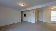 This single-floor residence offers a clean, functional layout designed for practical living. The main living area is spacious and open, featuring wall-to-wall neutral carpeting that adds a sense of warmth and continuity throughout the space. White walls and a flush ceiling light fixture keep the area feeling bright and airy.

A standout feature of the living area is a pass-through window cut into the interior wall, offering a convenient connection to an adjacent room — likely a kitchen — making the space feel interactive and well-connected. A louvered bi-fold closet door provides ample storage directly within the main living space, while a floor-level HVAC vent ensures year-round climate comfort.

The entryway is defined by a solid paneled front door that opens directly into the living area, creating an accessible and welcoming flow from the moment you step inside. A secondary doorway on the opposite side of the main wall leads further into the unit, suggesting additional rooms beyond the primary living space.

Overall, the unit presents a straightforward, no-frills design with a neutral color palette and functional features throughout, making it a versatile space well-suited for a variety of living arrangements.
