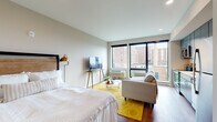 This well-appointed studio unit offers a thoughtfully designed open-concept layout that seamlessly integrates the living, sleeping, and kitchen areas into one cohesive space. Rich hardwood flooring flows throughout, grounding the neutral color palette and contributing to a warm, contemporary ambiance.

The kitchen is a standout feature, outfitted with sleek charcoal-toned cabinetry, a classic white subway tile backsplash, and a full suite of stainless steel appliances including a full-size refrigerator, electric range, over-the-range microwave, and dishwasher. Light countertops provide ample workspace, making the kitchen both functional and visually appealing.

The main living area accommodates a full sleeping arrangement alongside a comfortable seating area, offering versatility in how the space is used. A vibrant yellow area rug anchors the living zone, adding a pop of color against the otherwise understated décor. Recessed lighting throughout ensures even illumination across the entire unit.

Large, floor-to-ceiling style windows flood the space with natural light and frame views of the surrounding urban landscape. The bathroom features matching charcoal vanity cabinetry, a clean white countertop, and wood-look flooring that ties in with the rest of the unit. The overall design balances modern finishes with practical livability.