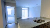 This single-floor space showcases an open-concept layout that seamlessly connects the kitchen and living area, creating a cohesive and functional environment. The kitchen is equipped with granite countertops featuring a speckled finish, complemented by a stainless steel sink and modern cabinetry. The countertop extends to form a peninsula, offering both additional workspace and a natural divider between the kitchen and the main living area.

The living area is characterized by clean, neutral-toned walls and smooth flooring, contributing to a bright and airy atmosphere. A large window fitted with vertical blinds dominates the far wall, allowing an abundance of natural light to fill the space while also providing access to what appears to be a balcony or outdoor area beyond. Recessed lighting in the ceiling supplements natural light, ensuring the space remains well-lit.

Crown molding runs along the ceiling perimeter, adding a refined architectural touch. A doorway visible to the left leads to an adjacent room, suggesting additional living or utility space. Overall, the unit presents a clean, modern aesthetic with practical design elements throughout.