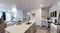 This single-floor residence combines modern design with functional living across a thoughtfully arranged floor plan. The open-concept kitchen serves as the heart of the home, anchored by a generously sized island with a curved edge, quartz countertops, and an undermount sink accented by globe pendant lighting. Gray shaker-style cabinetry lines the walls, complemented by a white subway tile backsplash and a full suite of stainless steel appliances, including a range, microwave, and refrigerator. Wood-look plank flooring flows seamlessly from the kitchen into the living area, where a ceiling fan, recessed lighting, and large windows with sliding glass door access to a private balcony invite natural light throughout. The bedrooms offer plush carpeting, generous window coverage with blinds and curtains, and ceiling fans for added comfort. One bedroom features a walk-in closet and direct access to additional storage. The bathroom is finished with gray cabinetry, a white quartz vanity top, wood-look flooring, and a tub-shower combination. Throughout the residence, crisp white walls and consistent finishes create a cohesive, contemporary aesthetic that feels both inviting and functional.