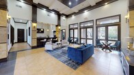 Located in Houston, TX, this single-floor lobby serves as a grand and welcoming communal space defined by its impressive architectural design. Soaring double-height ceilings anchored by dark wood-trimmed coffers and bold stone-clad columns immediately establish a sense of grandeur upon entry. The formal entryway features large floor-to-ceiling glass doors framed in dark wood, flooding the space with natural light and offering views of the surrounding exterior. An elegant chandelier hangs above the entry vestibule, complementing the warm tile flooring that flows throughout. The main lounge area is furnished with a plush navy velvet sofa, accent chairs, and a patterned area rug, creating an inviting setting for residents and guests. Curated artwork adorns the walls, adding character and depth to the neutral color palette of cream and warm gold tones. A built-in bar counter with stone-faced columns, pendant lighting, and bar seating provides a functional yet stylish social hub. Recessed lighting and wall sconces offer warm, layered illumination throughout. Additional seating areas with glass-top tables and contemporary chairs round out the space, while open sightlines between zones maintain a cohesive, airy atmosphere throughout this well-appointed lobby.