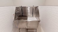 This single-level space is thoughtfully designed as a dedicated pet grooming room, combining functional equipment with a clean, easy-to-maintain aesthetic. The walls are clad in classic white subway tile from floor to mid-wall height, transitioning to a smooth painted finish above, creating a hygienic and visually tidy environment well-suited for its purpose.

The centerpiece of the room is a large stainless steel grooming tub, equipped with an integrated sprayer and restraint loops to safely secure pets during bathing. Under-counter cabinet storage is built into the tub station, offering convenient organization for grooming supplies. A non-slip grooming mat bearing a paw print motif is positioned nearby, providing a secure surface for pets during drying and styling.

A wall-mounted vacuum and blower unit is installed on the tiled wall alongside a coiled vacuum hose stored neatly on a wall hook, keeping the space organized and accessible. Electrical outlets and utility connections are thoughtfully integrated into the tile wall for convenient equipment use.

The room is accessed through a bold slate-blue five-panel door with brushed hardware, adding a touch of design character to an otherwise utilitarian space. Playful dog-themed artwork on the wall adds a welcoming, pet-friendly personality to the room's overall ambiance.
