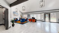 This single-story clubhouse facility is thoughtfully designed to serve as a dynamic community hub, combining functionality with bold contemporary style. The main lounge area immediately impresses with its soaring vaulted ceilings accented by rustic dark wood beams, marble-look tile flooring, and dramatic geometric chandelier lighting. Vibrant cobalt blue and burnt orange upholstered seating arrangements are anchored by a rich teal area rug, creating an energetic yet inviting atmosphere. A white brick fireplace adds a classic focal point, while large windows flood the space with abundant natural light and views of the surrounding landscape. A curved reception and bar-style counter, lined with navy velvet barstools on gold legs, provides a central gathering point and is adorned with curated contemporary artwork. A dedicated workstation alcove along one wall features built-in shelving and dual monitors, offering a convenient space for residents. Adjacent private offices provide additional functional areas. The fitness center is equally impressive, boasting vaulted ceilings, bold cobalt blue accent walls, floor-to-ceiling mirrors, and a comprehensive array of cardio and strength-training equipment, including cable machines, free weights, treadmills, and stationary bikes — all beneath a ceiling fan for added comfort.