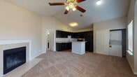 This single-story unit is defined by its open layout and vaulted ceilings, which lend a sense of spaciousness to the living area. A ceiling fan with light kit is centrally positioned beneath the pitched roofline, enhancing airflow and visibility throughout the main living space. A wood-surround fireplace serves as a focal point along one wall, providing both a functional and architectural accent.

The kitchen flows directly from the living area and features dark cabinetry contrasted by light countertops, a central island, and black appliances including a refrigerator, range, and microwave. The kitchen floor is finished in hardwood, transitioning to plush carpet in the living and bedroom areas. A closet adjacent to the kitchen provides additional storage, while an in-unit laundry area with a stacked washer and dryer is conveniently tucked nearby.

The bedrooms are carpeted and well-proportioned, each equipped with ceiling fans. One bedroom includes a sliding-door closet and a window with natural light. The bathroom features hardwood flooring, a long vanity with a stone-look countertop, dark cabinetry, a wide mirror, and medicine cabinets, offering ample storage and preparation space. Windows throughout the unit allow natural light to fill the interior.