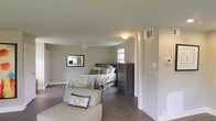 This single-floor space centers around a generously sized bedroom that combines comfort and practicality. The room is finished with warm hardwood-style flooring that contrasts cleanly against neutral gray walls, creating a calm and cohesive aesthetic throughout. A ceiling fan with a built-in light fixture is centrally positioned, providing both ventilation and ambient lighting, while a flush-mount ceiling light adds additional illumination. Multiple windows allow natural light to filter into the space, enhancing its open, airy feel. The bedroom accommodates a full-sized bed with a dark-toned headboard, complemented by a matching multi-drawer dresser that provides substantial storage. A framed wall mirror adds depth to the room, while a cushioned accent chair with decorative pillows offers a comfortable seating area. HVAC vents are visibly integrated into the walls, ensuring climate control. A partial view toward an adjacent area suggests additional rooms or utility spaces beyond. The overall design is understated and move-in ready, balancing functional storage and living space within a clean, modern aesthetic.