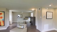 This single-floor space centers around a generously sized bedroom that combines comfort and practicality. The room is finished with warm hardwood-style flooring that contrasts cleanly against neutral gray walls, creating a calm and cohesive aesthetic throughout. A ceiling fan with a built-in light fixture is centrally positioned, providing both ventilation and ambient lighting, while a flush-mount ceiling light adds additional illumination. Multiple windows allow natural light to filter into the space, enhancing its open, airy feel. The bedroom accommodates a full-sized bed with a dark-toned headboard, complemented by a matching multi-drawer dresser that provides substantial storage. A framed wall mirror adds depth to the room, while a cushioned accent chair with decorative pillows offers a comfortable seating area. HVAC vents are visibly integrated into the walls, ensuring climate control. A partial view toward an adjacent area suggests additional rooms or utility spaces beyond. The overall design is understated and move-in ready, balancing functional storage and living space within a clean, modern aesthetic.