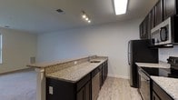 Located at 12001 Old Vine Blvd in Lewes, Delaware, this single-floor residence offers a clean, modern aesthetic throughout its thoughtfully designed layout. The kitchen is a standout feature, showcasing granite countertops with subtle speckled detailing that run along an extended peninsula, providing ample prep and dining space. Dark espresso cabinetry contrasts beautifully against the light-toned luxury vinyl plank flooring, which adds warmth and durability underfoot. Stainless steel appliances, including a refrigerator and dishwasher, complement the contemporary finish palette. A single-basin sink is neatly integrated into the countertop, accompanied by a sleek modern faucet. Track lighting and a flush skylight-style ceiling fixture provide bright, even illumination throughout the kitchen area. The light gray walls and white trim contribute to a crisp, neutral atmosphere that lends itself well to a variety of interior styles. The open feel of the space allows for natural flow between areas, making it well-suited for both daily living and entertaining. Overall, the residence presents a polished and move-in ready environment with modern upgrades throughout.