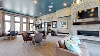 This single-story communal space is thoughtfully designed to blend sophisticated style with comfortable functionality. The entry hall sets a strong first impression with herringbone-patterned wood-look tile flooring, dark-trimmed doorways, and interior windows that offer glimpses into adjoining rooms, including a dedicated executive or private lounge area.

The main gathering room is a standout, anchored by a dramatic floor-to-ceiling stacked stone fireplace with a wall-mounted flat-screen television above. A teal-painted ceiling adds a bold, contemporary contrast to the neutral walls and carpet below, while globe-shaped gold pendant chandeliers provide warm, ambient lighting. Tall, dark-framed windows line the walls, flooding the space with natural light and views of the outdoors. Seating includes plush sofas, wingback chairs, and accent ottomans arranged around a rustic wooden coffee table.

Adjacent to the lounge is a dining area with a round table and upholstered chairs, positioned near a stylish wet bar featuring dark cabinetry, mosaic tile backsplash, granite countertops, a stainless steel sink, and a full-size refrigerator. A reading nook rounds out the space, showcasing a striking circular wall-mounted bookshelf paired with teal accent chairs, creating an inviting, intimate retreat within the larger layout.