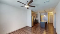 Located in Little Elm, this single-floor residence offers a thoughtfully designed open-concept layout that seamlessly connects the living and kitchen areas. Rich, dark hardwood-style flooring flows throughout the space, adding warmth and continuity to the interior. The living area is appointed with a contemporary ceiling fan, contributing both style and comfort to the room.

The kitchen is a standout feature, showcasing a full suite of stainless steel appliances, including a refrigerator, range, and built-in microwave. Medium-toned cabinetry provides ample storage, while a kitchen island topped with a light granite-style countertop serves as both a functional workspace and a casual gathering spot. Pendant lighting above the island adds a refined finishing touch.

Track lighting installed along the ceiling further enhances the modern aesthetic of the space, ensuring well-distributed illumination throughout. The clean white walls provide a neutral backdrop that complements the warm flooring and cabinetry tones. The open flow between the living and kitchen areas creates a spacious, airy feel, making the residence well-suited for both everyday living and entertaining. Overall, the space presents a polished, contemporary character with practical, high-quality finishes.