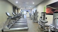 This fitness center offers a spacious, single-floor layout designed to accommodate a wide variety of exercise needs. The cardio section is prominently featured along one side of the room, lined with a row of True-brand treadmills equipped with individual consoles, providing ample space for cardio workouts. Opposite the treadmills, a selection of cable and resistance strength-training machines are arranged to allow users to move efficiently between exercises. The flooring consists of durable, textured material suited for high-traffic gym use, providing both safety and stability underfoot. Overhead, recessed lighting and ceiling-mounted fixtures illuminate the space evenly, creating a bright and energizing atmosphere. A ceiling fan toward the rear of the room aids in air circulation. The walls are finished in neutral tones, contributing to a clean and focused environment. Toward the back of the facility, additional equipment and what appears to be a reception or utility area can be seen, suggesting further amenities beyond the main workout floor. A wall-mounted television is also visible, adding an element of entertainment during workouts.