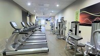 This fitness center offers a spacious, single-floor layout designed to accommodate a wide variety of exercise needs. The cardio section is prominently featured along one side of the room, lined with a row of True-brand treadmills equipped with individual consoles, providing ample space for cardio workouts. Opposite the treadmills, a selection of cable and resistance strength-training machines are arranged to allow users to move efficiently between exercises. The flooring consists of durable, textured material suited for high-traffic gym use, providing both safety and stability underfoot. Overhead, recessed lighting and ceiling-mounted fixtures illuminate the space evenly, creating a bright and energizing atmosphere. A ceiling fan toward the rear of the room aids in air circulation. The walls are finished in neutral tones, contributing to a clean and focused environment. Toward the back of the facility, additional equipment and what appears to be a reception or utility area can be seen, suggesting further amenities beyond the main workout floor. A wall-mounted television is also visible, adding an element of entertainment during workouts.