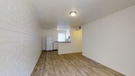 This single-floor space features an open-concept design that combines the living area and kitchen into one cohesive layout. The flooring throughout is light wood-style laminate, lending a clean and modern feel to the interior. A notable architectural detail is the exposed brick accent wall along one side of the living area, adding texture and visual interest to the otherwise neutral, white-painted walls.

The kitchen is positioned toward the rear of the space and is partially separated by a peninsula counter with a dark wood-toned surface, providing both a functional workspace and a natural divider between the cooking and living zones. White appliances, including a refrigerator and what appears to be a dishwasher or range, are integrated into the kitchen layout. A window above the kitchen counter allows natural light to filter into the area, enhanced by vertical blinds for light control.

Flush-mounted ceiling light fixtures illuminate both the living and kitchen areas. A doorway visible at the rear of the space leads to an additional room or hallway. The overall ambiance is simple, bright, and functional, with a neutral palette that offers versatility for various furnishing styles.