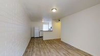 This single-floor space features an open-concept design that combines the living area and kitchen into one cohesive layout. The flooring throughout is light wood-style laminate, lending a clean and modern feel to the interior. A notable architectural detail is the exposed brick accent wall along one side of the living area, adding texture and visual interest to the otherwise neutral, white-painted walls.

The kitchen is positioned toward the rear of the space and is partially separated by a peninsula counter with a dark wood-toned surface, providing both a functional workspace and a natural divider between the cooking and living zones. White appliances, including a refrigerator and what appears to be a dishwasher or range, are integrated into the kitchen layout. A window above the kitchen counter allows natural light to filter into the area, enhanced by vertical blinds for light control.

Flush-mounted ceiling light fixtures illuminate both the living and kitchen areas. A doorway visible at the rear of the space leads to an additional room or hallway. The overall ambiance is simple, bright, and functional, with a neutral palette that offers versatility for various furnishing styles.