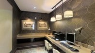 This single-floor space functions as a well-appointed workspace designed with both style and productivity in mind. The room is defined by its striking dark wallpaper, which features a bold geometric ogee pattern with subtle metallic accents, creating a rich and refined atmosphere throughout.

A continuous L-shaped built-in desk wraps along two walls, providing generous surface area for multiple workstations. The desk accommodates computers, a printer, and additional equipment, making it highly functional for professional or administrative use. Beneath the desk, open shelving offers practical storage for books and files.

Lighting is a key feature of this space. A track-mounted pendant system with three large drum shades hangs above the primary work surface, delivering focused, warm illumination. This is complemented by wall-mounted picture lights that highlight two framed architectural prints, adding a curated, gallery-like quality to the room.

The ceiling is clean and white, contrasting elegantly with the darker walls and grounding the overall design. The floor features a decorative area rug that adds texture and warmth. The overall ambiance is polished and professional, balancing decorative sophistication with practical workspace functionality.