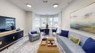 This inviting social room in Lakeland, Tennessee, is a well-appointed communal gathering space designed for relaxation and casual socializing. The room features warm hardwood-style flooring throughout, neutral light gray walls, and flush-mount ceiling lighting that keeps the space bright and welcoming.

The main lounge area is anchored by a plush blue sofa and coordinating upholstered armchairs arranged around a woven coffee table atop a patterned area rug. Board games, including chess and backgammon sets, are displayed on the coffee table, adding a recreational touch. A sleek media console with decorative detailing supports a wall-mounted flat-screen television along one wall, while framed artwork adds a tasteful finishing accent.

A distinctive bay window nook at one end of the room floods the space with natural light and offers views of the tree-lined surroundings outside. A round dining table with modern navy chairs is positioned within this nook, creating an intimate spot for small gatherings or casual conversation.

A set of glass-paneled double doors connects the social room to an adjoining fitness area, providing easy access and visual openness between the two spaces. The overall ambiance is modern, comfortable, and functional — ideal for community interaction and leisure.