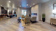 Located at 9110 Crown Springs View in Colorado Springs, Colorado, this single-floor space is thoughtfully designed with a contemporary aesthetic that blends comfort and functionality throughout. Light-toned luxury vinyl plank flooring runs continuously across the entire space, creating a cohesive and clean foundation that complements the neutral gray and white color palette.

The primary lounge area serves as a focal point of the space, anchored by a dramatic floor-to-ceiling textured stone accent wall housing a sleek, linear electric fireplace with vibrant blue and purple flames. Patterned accent chairs and a leather bench are arranged around a wood coffee table, offering an inviting seating arrangement ideal for gathering. Recessed lighting illuminates the area throughout.

A secondary lounge area provides additional seating with leather chairs and sofas arranged around a wall-mounted television set against a wood-paneled backdrop. Tall floor lamps and curated décor contribute to the relaxed, welcoming atmosphere of this open zone.

The kitchen area is well-appointed with dark cabinetry, light quartz countertops, stainless steel appliances, and a commercial-style beverage station. A generous bar-height island with seating for multiple guests anchors the space, lit by a row of modern pendant lights. Large windows allow natural light to filter in, connecting the interior to the outdoor surroundings.