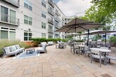 This well-appointed outdoor courtyard at 9142 Richmond Hwy in Fort Belvoir, Virginia, is nestled within the interior of a contemporary multi-story building complex featuring a modern facade of gray paneling, large windows, and private balconies. The courtyard is thoughtfully designed to offer a variety of social and recreational areas across its expansive paved grounds.

A standout feature is the oversized outdoor chess set, positioned on a dedicated mat surface, providing an engaging focal point and activity space for residents and visitors. Nearby, a striking sculptural tree installation adds an artistic element to the outdoor environment.

The courtyard is divided into distinct zones: a relaxed lounge area outfitted with comfortable upholstered sofas and low coffee tables, ideal for casual gatherings, and a more structured outdoor dining area furnished with multiple round and rectangular tables paired with chairs and large canopy umbrellas for shade. Warm-toned brick paver walkways and open stone tile plazas connect these spaces seamlessly.

Lush landscaping frames the perimeter, with mature trees, flowering shrubs, ornamental grasses, and colorful plantings providing greenery and privacy. Decorative lighting poles are positioned throughout for evening ambiance. The enclosed nature of the courtyard creates a sheltered, community-centered atmosphere that encourages outdoor living and social interaction year-round.