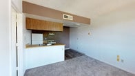 This single-floor unit offers a functional and straightforward layout designed for comfortable everyday living. The open-concept kitchen and living area form the heart of the space, with the kitchen featuring warm wood-toned cabinetry both above and below the counters, a double-basin stainless steel sink set into a light laminate countertop, a white electric range with coil burners, a white refrigerator, and an overhead ventilation hood. The kitchen is defined by a peninsula that separates it from the adjacent living area, which is finished with plush carpet and neutral-toned walls. Dark wood-look vinyl flooring runs throughout the kitchen, providing a durable and easy-to-clean surface. A textured popcorn ceiling and ceiling-mounted HVAC vents are consistent throughout the main living space. The bedroom is a quiet, well-proportioned room with soft carpeting, clean white walls, and a window that invites natural light and a view of surrounding greenery. The full bathroom includes a soaking tub with a tiled surround featuring a frosted tile pattern, a single vanity sink with wood cabinetry beneath, a wall-mounted mirror, and dark wood-look vinyl flooring that ties back to the kitchen aesthetic. Overall, the unit presents a clean, no-frills interior with practical finishes throughout.