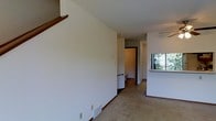 This two-story residential space offers a straightforward and functional layout designed for comfortable everyday living. The main level features open living areas finished with wall-to-wall carpeting and clean white walls that lend a bright, neutral ambiance throughout. A staircase with a warm wood handrail connects the two floors, serving as a central architectural element visible from the main living area. The kitchen is situated adjacent to the living space and includes a pass-through counter opening that maintains visual connectivity between the rooms. A window in the kitchen area allows natural light to filter in, offering a pleasant view of surrounding greenery. The lower level also features access to additional rooms, including what appears to be a bathroom, accessible through a doorway visible from the main hall. The overall design is clean and unadorned, prioritizing utility and livability. Neutral tones, consistent flooring, and straightforward room transitions create a cohesive and easy-to-navigate environment. The second floor, accessible via the interior staircase, expands the overall living capacity of the space, making it well-suited for a variety of occupancy needs.