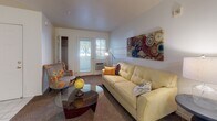 This single-floor residence offers a practical and cohesive layout designed for comfortable everyday living. The living room serves as the central gathering space, featuring warm carpeted flooring, a tufted sofa, an accent chair, and a glass-top coffee table. A patio door allows natural light to filter in and provides direct outdoor access. The room transitions smoothly into the kitchen area via tile flooring, creating a clear distinction between spaces.

The kitchen is efficiently arranged with light wood cabinetry extending to the upper walls, white appliances including a refrigerator, electric range, and over-the-range microwave, and a clean white countertop offering ample prep space. The tile flooring continues throughout, lending a bright, easy-to-maintain quality.

The bedroom is a quiet, carpeted retreat equipped with a ceiling fan, a window with blinds for light control, and sufficient space for a full-sized bed and furnishings. The bathroom features a full tub-shower combination with a curved curtain rod, light wood vanity cabinetry, a white countertop, and tile flooring. The overall aesthetic throughout the unit is neutral and clean, with white walls providing a versatile backdrop.
