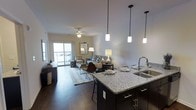 This single-floor residence showcases a thoughtfully designed open-concept layout that seamlessly connects the kitchen and living areas, creating a spacious and inviting atmosphere. The kitchen is a standout feature, centered around a large granite-topped island with seating and a double stainless steel sink. Dark cabinetry with brushed hardware adds a sophisticated contrast to the light stone countertops, while three elegant pendant lights hang overhead, providing both functional and ambient illumination. Rich, dark hardwood flooring flows throughout the entire living space, grounding the design with warmth and continuity. The living area is generously proportioned, offering ample room for comfortable furnishings and social gatherings. A ceiling fan overhead ensures year-round comfort, while a sliding glass door at the far end of the room floods the interior with natural light and provides direct access to an outdoor balcony, extending the living space outdoors. The neutral color palette of warm whites and grays throughout creates a clean, modern aesthetic. Overall, the space balances contemporary finishes with a welcoming ambiance, making it both functional and visually appealing.