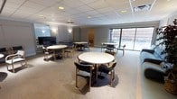 This single-floor multipurpose gathering space is designed to accommodate group meetings, social events, and communal activities. The main room is generously proportioned, featuring multiple round white tables surrounded by wood-framed chairs with padded gray upholstery, arranged throughout the open floor plan to encourage conversation and collaboration. The drop ceiling with recessed lighting and wall-mounted sconces provides warm, even illumination throughout the space. Soft neutral-toned carpet adds comfort underfoot and contributes to a welcoming ambiance. Large windows along one wall flood the room with natural light and offer views of an adjacent outdoor patio area, creating a pleasant indoor-outdoor connection. A flat-screen television is mounted along one wall, making the space well-suited for presentations or group viewings. At the rear of the main room, a dedicated kitchenette is equipped with cabinetry, a refrigerator, and a countertop workspace with a range hood, providing convenient support for catering and refreshments during events. The overall layout is practical and flexible, making this space highly functional for a wide range of uses including community gatherings, meetings, dining, and social programming.