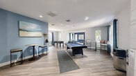 This single-floor game and recreation room offers a well-appointed space designed for entertainment and socializing. The open layout is finished with wood-style luxury vinyl flooring throughout, complemented by crisp white ceilings with recessed lighting that ensures the space is well-lit at all times. The walls feature a thoughtful combination of light gray and bold navy blue accent walls, lending the room a modern, energetic character.

At the heart of the space sits a full-size pool table with a blue felt surface, positioned atop a gray area rug. Adjacent to it, a foosball table provides additional gaming options. A dedicated lounge alcove features comfortable navy blue sofas and armchairs arranged around a glass coffee table, with framed wall art and large windows dressed in teal curtain panels that allow natural light to flow in and offer views of the surrounding outdoors.

Classic arcade cabinets, including titles such as X-Men and Mortal Kombat, are positioned along one wall, adding a nostalgic touch to the entertainment offerings. High-top bistro tables and bar stools are distributed throughout, providing casual seating for spectators and guests. A water cooler and waste station are conveniently located along one wall. Multiple windows throughout the room ensure abundant natural light and a welcoming, open ambiance.
