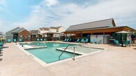 This outdoor amenity area is centered around a generously sized swimming pool with clear, blue-green water. The pool includes built-in in-water lounge chairs, allowing residents to relax partially submerged, and is accompanied by an adjacent raised spa for additional leisure options. The surrounding pool deck is finished in a warm, neutral concrete surface that provides ample space for lounging and circulation.

Numerous teal-colored lounge chairs are arranged along all sides of the pool deck, complemented by shaded umbrella stations that offer relief from direct sunlight. A wrought-iron perimeter fence encloses the entire pool area, providing both security and a clean visual boundary.

Adjacent to the pool, a single-story clubhouse structure serves as a functional anchor to the space. The building features natural wood cladding, a standing-seam metal roof, and a covered outdoor porch equipped with ceiling fans and pendant lighting. This shaded overhang extends the usable space and provides a comfortable retreat from the open sun.

The overall ambiance is modern yet approachable, with consistent teal accents throughout the furniture tying the design together. Residential buildings visible beyond the fence provide a neighborhood backdrop to this well-appointed community amenity space.
