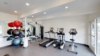 The fitness center at 2799 Adrian in San Diego is a well-appointed, single-floor facility designed to support a well-rounded workout routine. The space features crisp white walls and a neutral grey tile floor, creating a clean and modern aesthetic throughout. Recessed ceiling lighting provides bright, even illumination across the entire area, contributing to an energetic and focused atmosphere.

The cardio section is equipped with a treadmill, an elliptical trainer, and a stationary upright bike, offering a variety of options for cardiovascular exercise. A multi-function cable and weight machine anchors the strength training area, complemented by a flat workout bench and a collection of free weights and accessories along the wall. A wall-mounted air conditioning unit ensures comfortable temperatures year-round.

A large mirror spanning one wall enhances the sense of space, allows users to monitor their form during workouts, and reflects the equipment and natural brightness of the room. The overall layout is thoughtfully organized, keeping cardio and strength zones distinct while maintaining an open, uncluttered feel that accommodates multiple users at once.