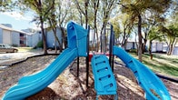 This outdoor playground is a well-appointed recreational space situated within a residential community, surrounded by a canopy of mature trees that provide natural shade throughout the area. The ground is covered with a wood chip surface, offering a safe and natural-looking base beneath the equipment.

The centerpiece of the playground is a vibrant blue multi-activity play structure, which features two slides — including a wide curved slide — a step ladder for easy access, and an enclosed tunnel element, making it an engaging destination for younger children. Adjacent to this main structure is a separate set of metal play equipment finished in black, featuring a red slide, railed ramps, and platform sections suited for older, more active children.

Adding further variety, the playground includes a spinning merry-go-round style apparatus and a set of chain swings, offering a diverse range of physical activities and play opportunities. The equipment is constructed with durable steel frames, designed to withstand outdoor conditions.

The surrounding environment features open green lawn areas, paved walkways, and views of neighboring residential buildings, contributing to a welcoming community atmosphere. The tree coverage ensures the space remains pleasant throughout different times of day, making this playground an inviting and functional outdoor amenity for residents of all ages.