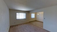 This single-floor residence offers a straightforward and functional layout designed for comfortable everyday living. The main living area is finished with plush wall-to-wall carpeting and is bathed in natural light through large windows dressed with vertical blinds, creating a bright and welcoming atmosphere. The space flows seamlessly into an adjoining dining area, which transitions to easy-care tile flooring — a practical and clean design choice that defines the two zones without the need for walls or partitions. A classic pendant light fixture hangs above the dining area, providing focused overhead illumination and adding a subtle decorative touch. The walls throughout are finished in a neutral, warm tone that complements both the carpeted and tiled sections of the floor plan. The open-concept connection between the living and dining spaces enhances the sense of roominess and allows for flexible furniture arrangements. Baseboards and clean wall lines contribute to the tidy, well-maintained character of the interior. Overall, this residence presents a clean, move-in-ready environment with a simple, cohesive aesthetic suited to a wide range of lifestyles.